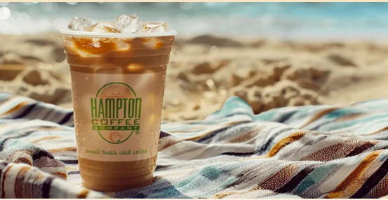 Iced Hamptons Coffee drink sitting on a beach blanket with sand and ocean in the background.